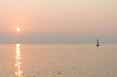 A sail boat peacefully sails at the horizon at suset time
