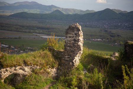 Ancient stone ruins overlook a vibrant valley with lush green hills and distant mountains under a warm sunset glow.の写真素材