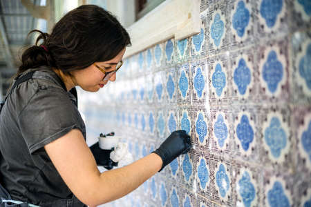 Faro, Portugal - April 5, 2021: Qualified woman cleaning and repairing tiles on the facade of old...