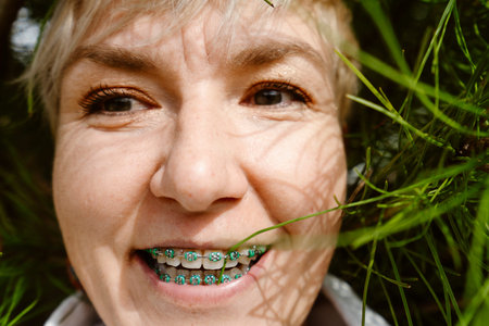 Portrait of a happy woman smiling with orthodontic braces while enjoying nature. Young adult female with dental braces outdoors among trees and flowers, expressing confidence, joy and positive lifestyle.の写真素材