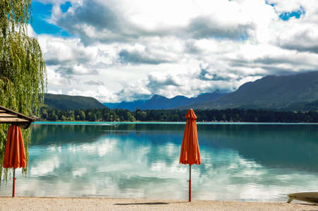 Empty beach bar on a mountain lake, view over the water with cloud reflections, two picturesque parasols in the foregroundの写真素材