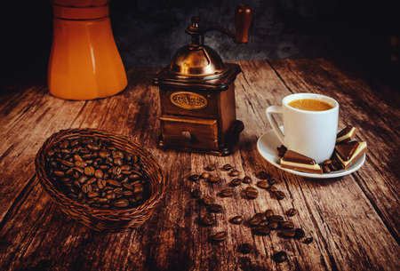 roasted coffee beans in a basket with an antique metal and wooden hand coffee grinder and a white cup with chocolates on wood with an orange Italian coffee pot in the dark backgroundの写真素材