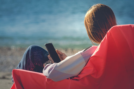 Young woman sitting on the beach and using a smart phone in the morningの写真素材