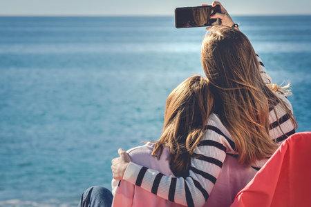 Back view of mother and daughter taking selfie with smartphone on the beach