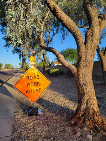 Bright diamond-shaped traffic sign with a light on its top set under eucalyptus tree on the verge of the walkway notifying about Road Work Ahead, Phoenix, Arizonaの写真素材