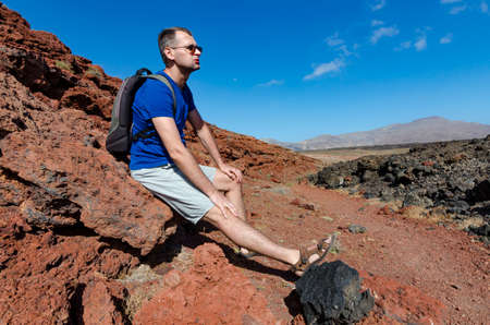 A man with a backpack sitting on a red rock and resting while trekking in volcanic landscape of...