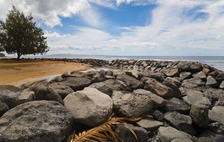 Tropical scene of a rock jetty enclosing a small lagoon by the ocean. Large tree in the background, wispy clouds and blue sky above, palm frond on the rocks in foreground.の写真素材