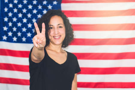 Young woman with the United States flag in the background celebrating and making victory with her handの写真素材