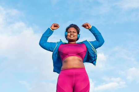 happy and excited african woman celebrating victory , overcoming challenges, body positive, exercising outside. Listening to music through earphones. High quality photoの写真素材