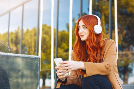 Young redhead woman sitting while listening to music with wireless headphones and smartphone,. High...
