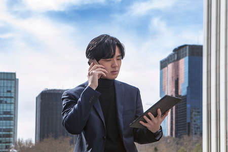 asian man outside the business center, talking on his cell phone, while looking at his tablet. High quality photoの写真素材