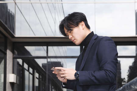 asian man outside the workplace looking at his mobile phone, glass building background . High quality photoの写真素材