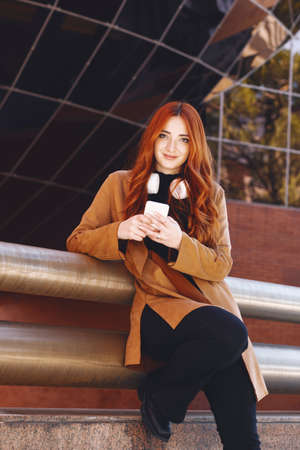 young red-haired girl looking into the camera, with wireless headphones and holding her cell phone.