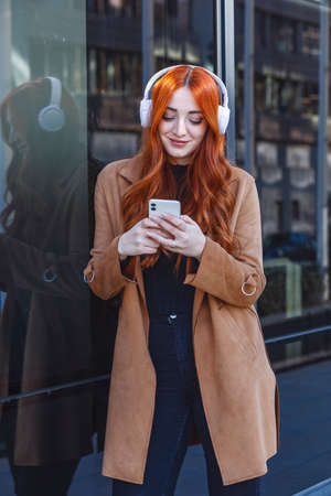 Young red-haired woman with wireless headphones looking at her cell phone while leaning against a wall. High quality photoの写真素材
