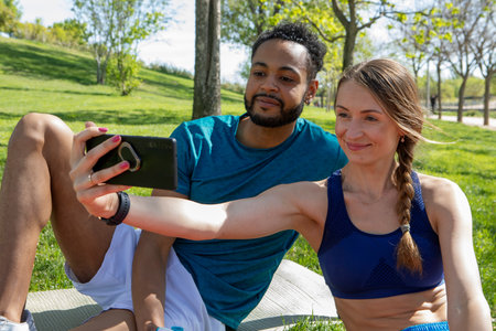 young multiethnic couple, dressed in sportswear, taking photos with their mobile phone, while taking...