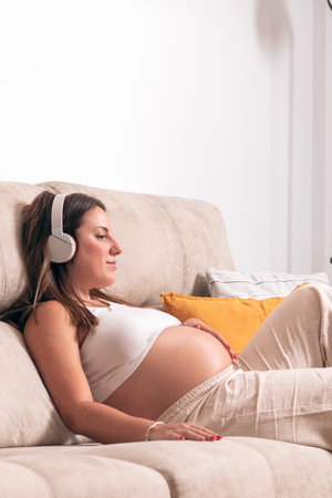 Young pregnant woman listening to music with headphones while relaxing on the couch at home. High quality photoの写真素材