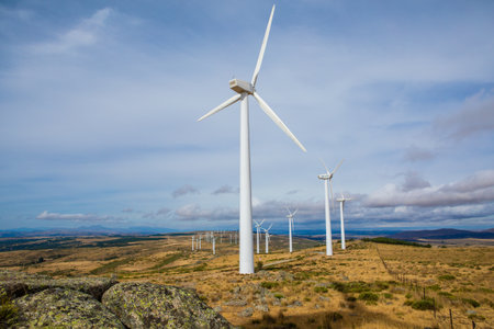 Tall wind turbines stand in a beautiful landscape, surrounded by rolling hills. The road meanders...
