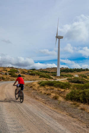 A person in a red shirt cycles on a gravel path surrounded by grass and hills, with a wind turbine visible nearby against a backdrop of blue and white clouds.の写真素材
