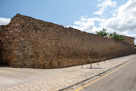Historic stone wall stretches along the side of a cobblestone street, showcasing ancient craftsmanship under a sunny sky with clouds. Nearby greenery adds to the scenery.の写真素材