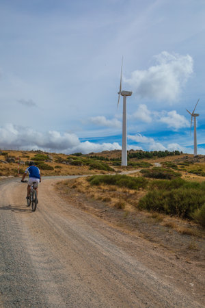 A cyclist rides along a dirt road surrounded by wind turbines. The landscape features rolling hills and a mix of shrubbery under a partly cloudy sky, creating a serene atmosphere.の写真素材