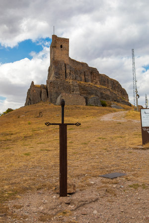 Visitors explore a striking castle ruin atop a rocky hill in Atienza, Guadalajara, Spain, with a prominent sword monument in the foreground under a cloudy sky.の写真素材