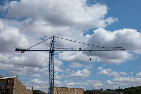A construction crane rises above a building site under a bright sky filled with fluffy white clouds....