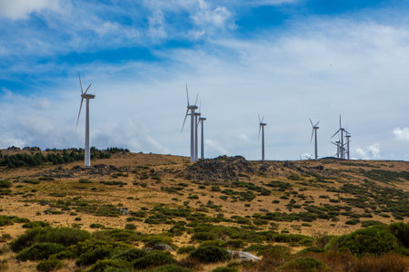 Tall wind turbines stand in a beautiful landscape, surrounded by rolling hills. The road meanders through the area under bright blue skies and fluffy white clouds, showing renewable energy.の写真素材