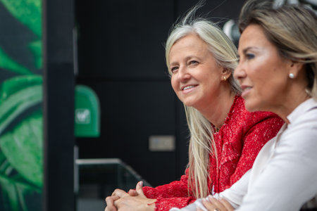 Two women are engaged in a lively conversation while reading on a glass railing. The setting features vibrant green decor, creating a cheerful atmosphere.の写真素材