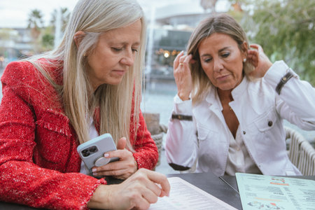 Two women sit at a table in a cafe, intently reviewing the menu. The setting has a relaxed atmosphere with natural light and views of the waterfront.の写真素材