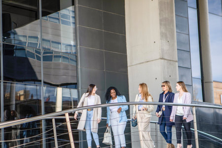 Five women dressed in stylish outfits walk together down a staircase in front of a contemporary building on a bright day, enjoying each other's company and the atmosphere.の写真素材