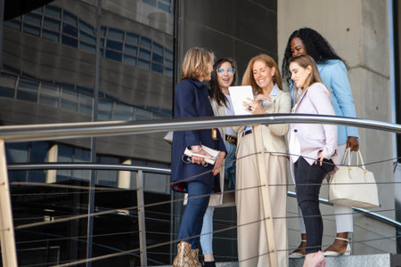 A group of five women engaged in a discussion at a contemporary building's balcony on a sunny afternoon. They are stylishly dressed and appear to be sharing ideas.の写真素材