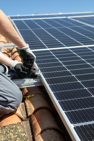 A worker in safety gear is carefully installing solar panels on a rooftop under bright blue skies. The task involves securing the panels firmly to ensure efficiency and stability.の写真素材