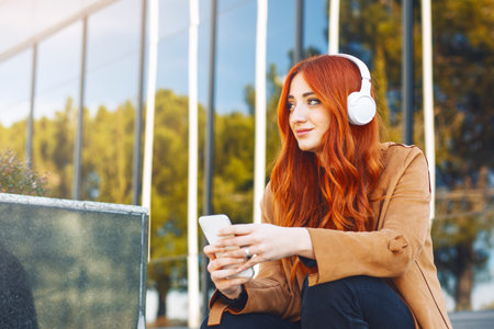 Young redhead woman sitting while listening to music with wireless headphones and smartphone,. High quality photoの写真素材