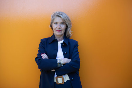 A confident woman with blonde hair stands against a vibrant orange wall. She appears self-assured with her arms crossed and a friendly expression on her face.の写真素材