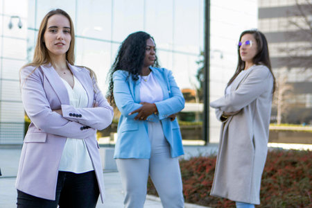 Three women with diverse styles pose confidently outside a sleek office structure. The sun shines brightly as they show strength and camaraderie in their business attire.の写真素材