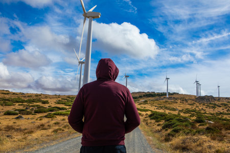 A person admires towering wind turbines against a backdrop of rolling terrain and dynamic clouds. The atmosphere is peaceful, inviting reflection on sustainable energy.の写真素材