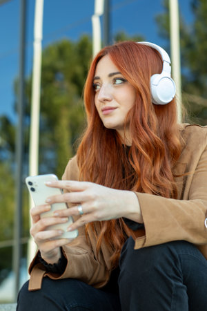 A young woman with flowing red hair sits outdoors, wearing headphones. She holds a smartphone and looks thoughtfully at her surroundings, enjoying the nice weather.の写真素材