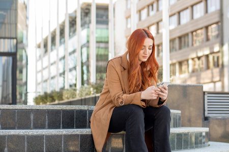 A young woman with red hair sits on stone steps, focused on her phone. The modern building reflects sunlight, creating a lively urban atmosphere.の写真素材