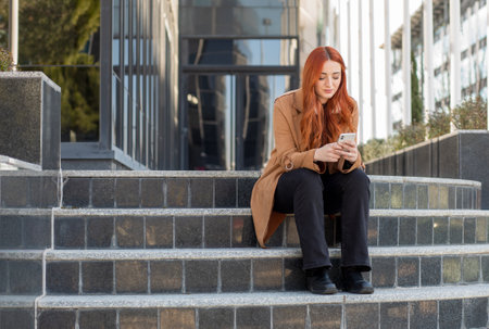 A young woman with red hair sits on stone steps, focused on her phone. The modern building reflects sunlight, creating a lively urban atmosphere.の写真素材