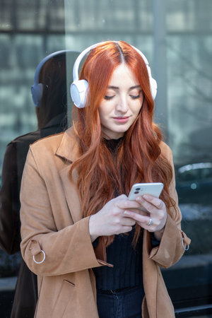 A young woman with long red hair enjoys music on her phone while wearing stylish headphones. She stands in front of a glass building, smiling at the camera.の写真素材