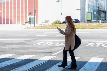 A woman with long red hair walks across a crosswalk in a modern city, focused on her smartphone. The weather is clear and sunny, highlighting the urban environment.の写真素材