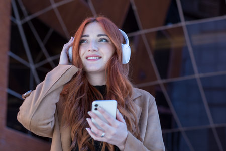 A woman with long red hair smiles while listening to music outdoors. She wears white headphones and...