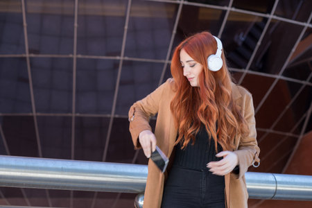 A young woman with vibrant red hair listens to music with white headphones while standing on a balcony. The modern architecture creates a stylish background.の写真素材