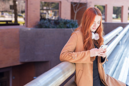 A young woman with long red hair is engaged with her smartphone, leaning casually on a railing in a stylish urban area during daylight. She appears relaxed and focused.の写真素材