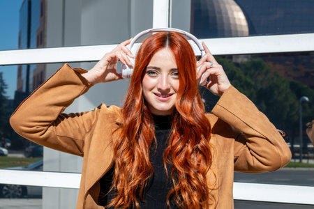 A woman with long red hair smiles joyfully while listening to music on headphones. She is dressed in...