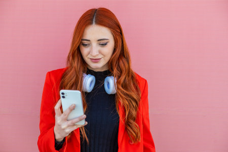 A young woman with long red hair checks her smartphone while wearing headphones. She stands against a striking pink wall, showing off a stylish outfit.の写真素材