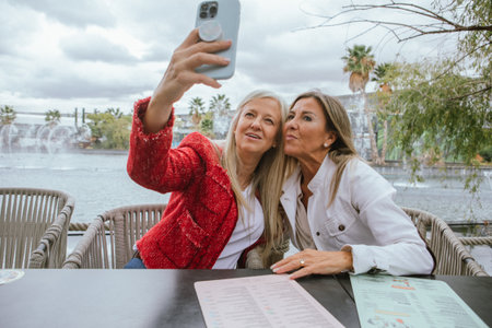 Two friends take a cheerful selfie together while sitting at a table by a beautiful fountain. The...