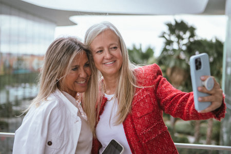 Two friends pose together for a selfie at a contemporary shopping center filled with greenery. They smile and enjoy their time while capturing the moment.の写真素材