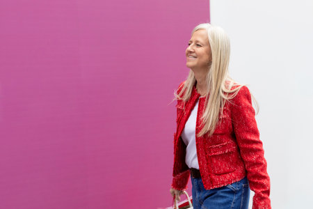 A woman with long, straight blonde hair is smiling as she walks confidently down the street. She wears a bright red jacket and casual jeans, creating a cheerful scene.の写真素材