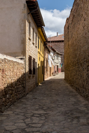 A narrow cobblestone street features stone walls and four buildings, with laundry hanging outside. The scene represents a peaceful moment in a preserved village under blue skies.の写真素材